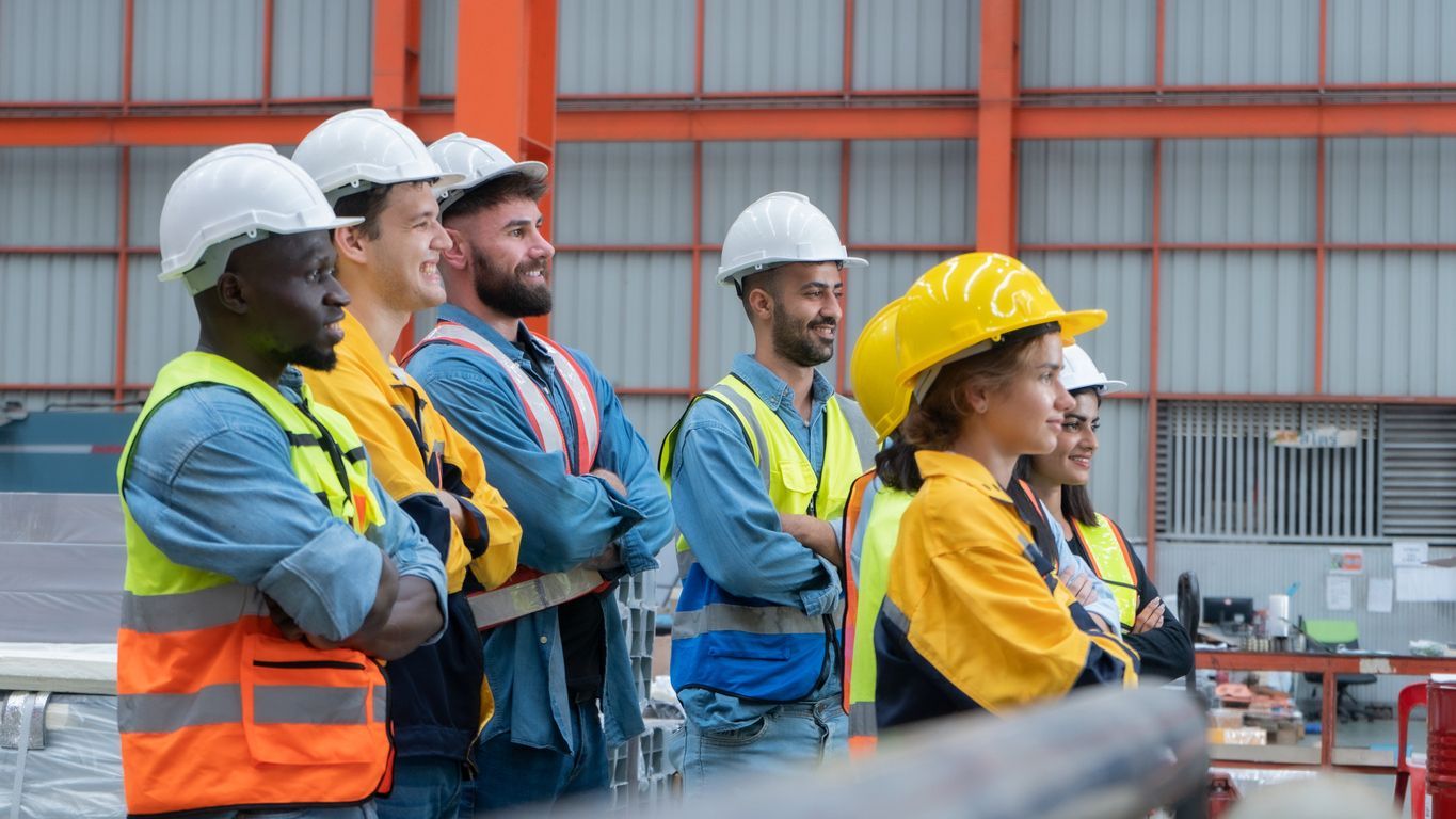 Workers in hard hats and safety vests standing together in an industrial warehouse, listening attentively.