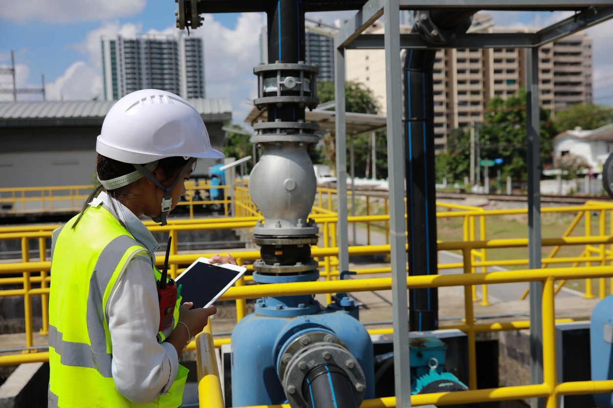 Worker in white hard hat and yellow vest inspecting industrial piping at a plant.