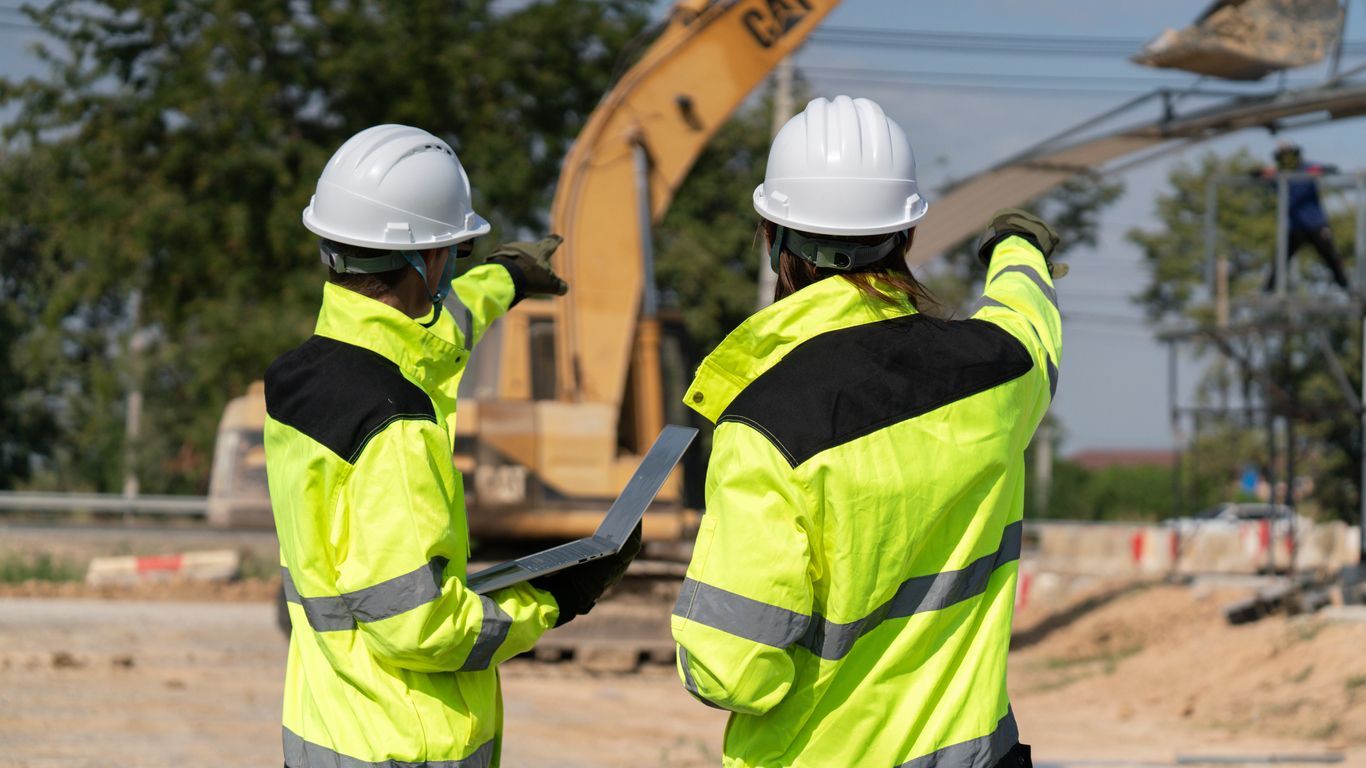 Two construction workers in hard hats and safety vests discussing on a job site with heavy equipment nearby
