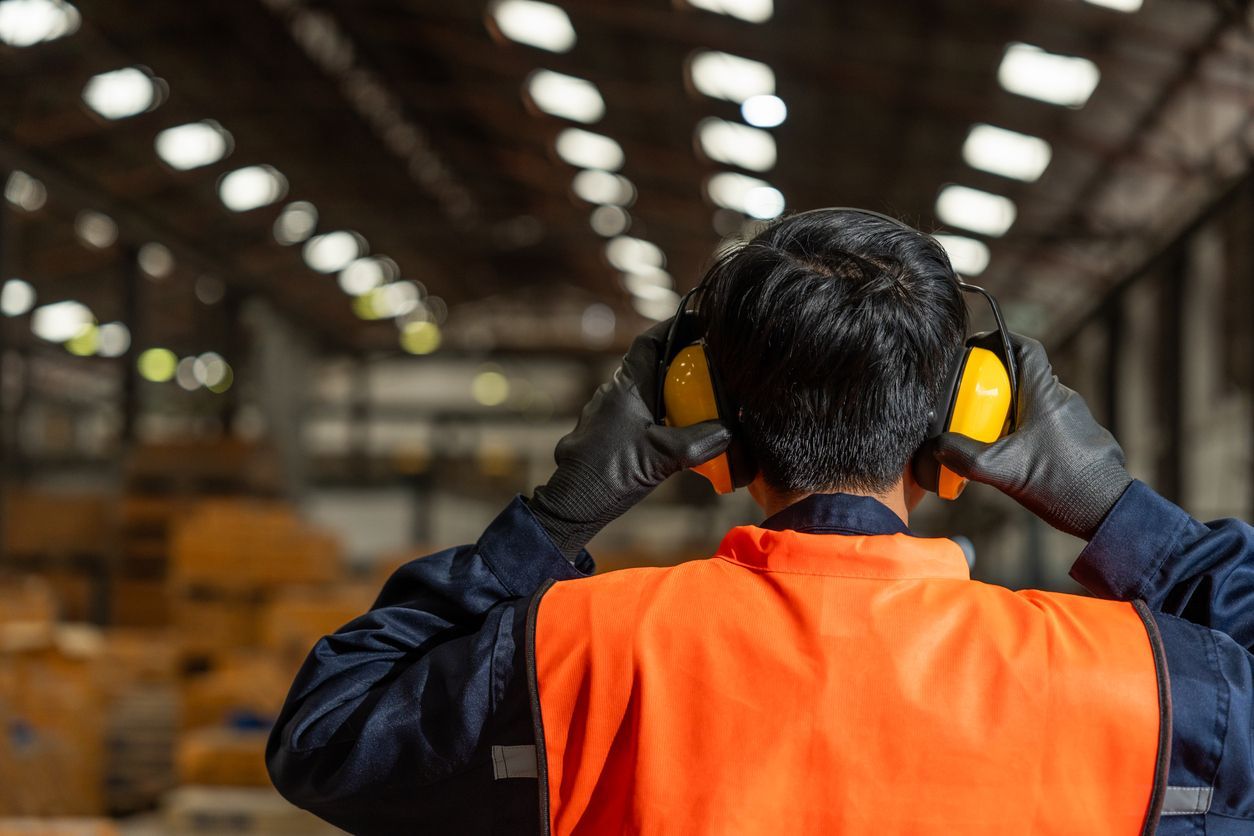 Warehouse worker in orange safety vest and yellow ear protection facing stacked boxes in a storage aisle