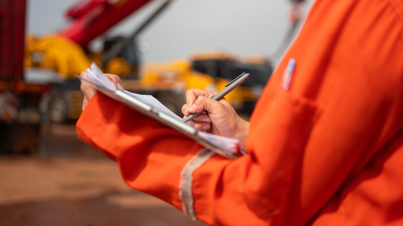 Person in orange safety gear writing on a clipboard at an outdoor work site