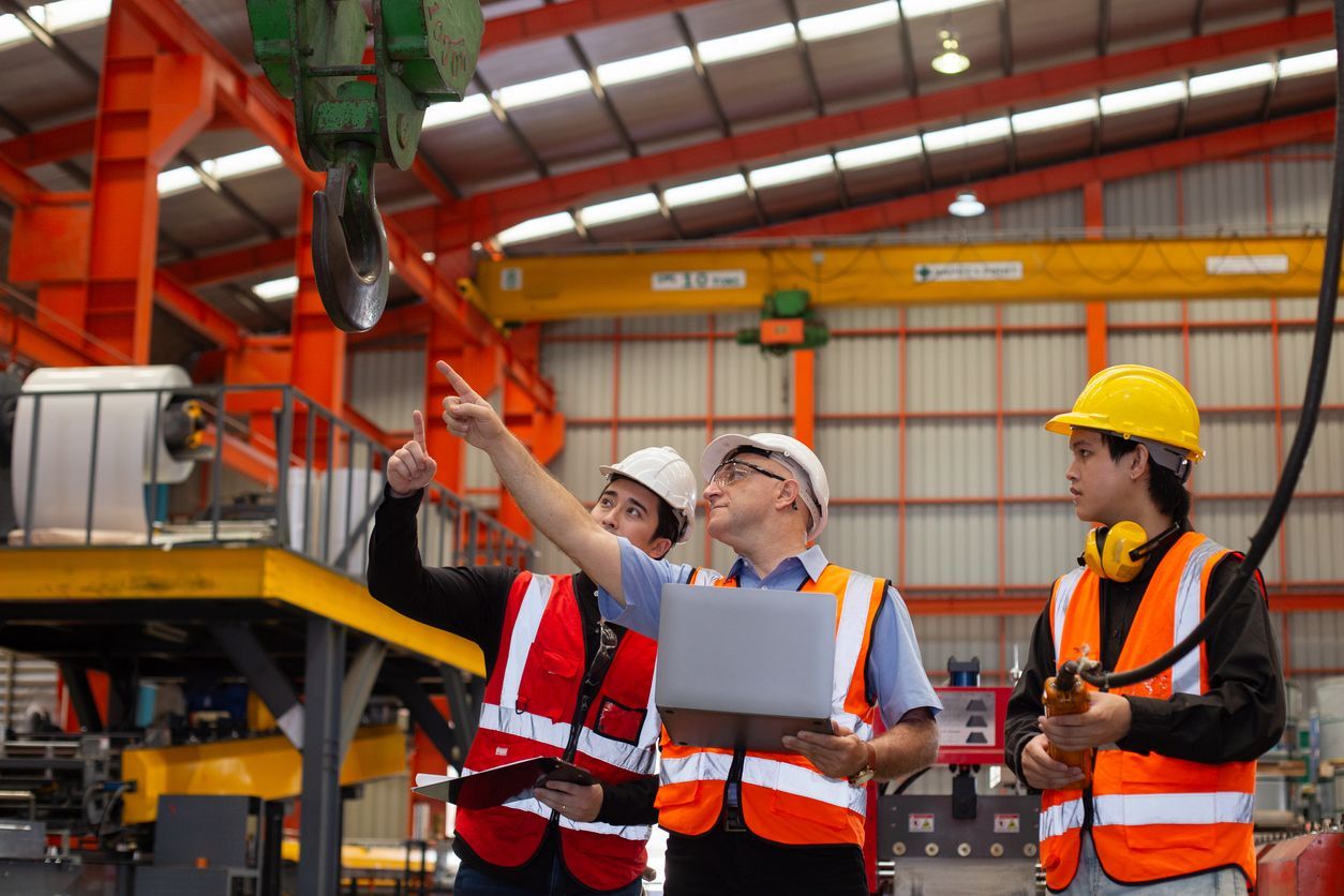 Three workers in safety vests and hard hats inspect machinery in a factory.