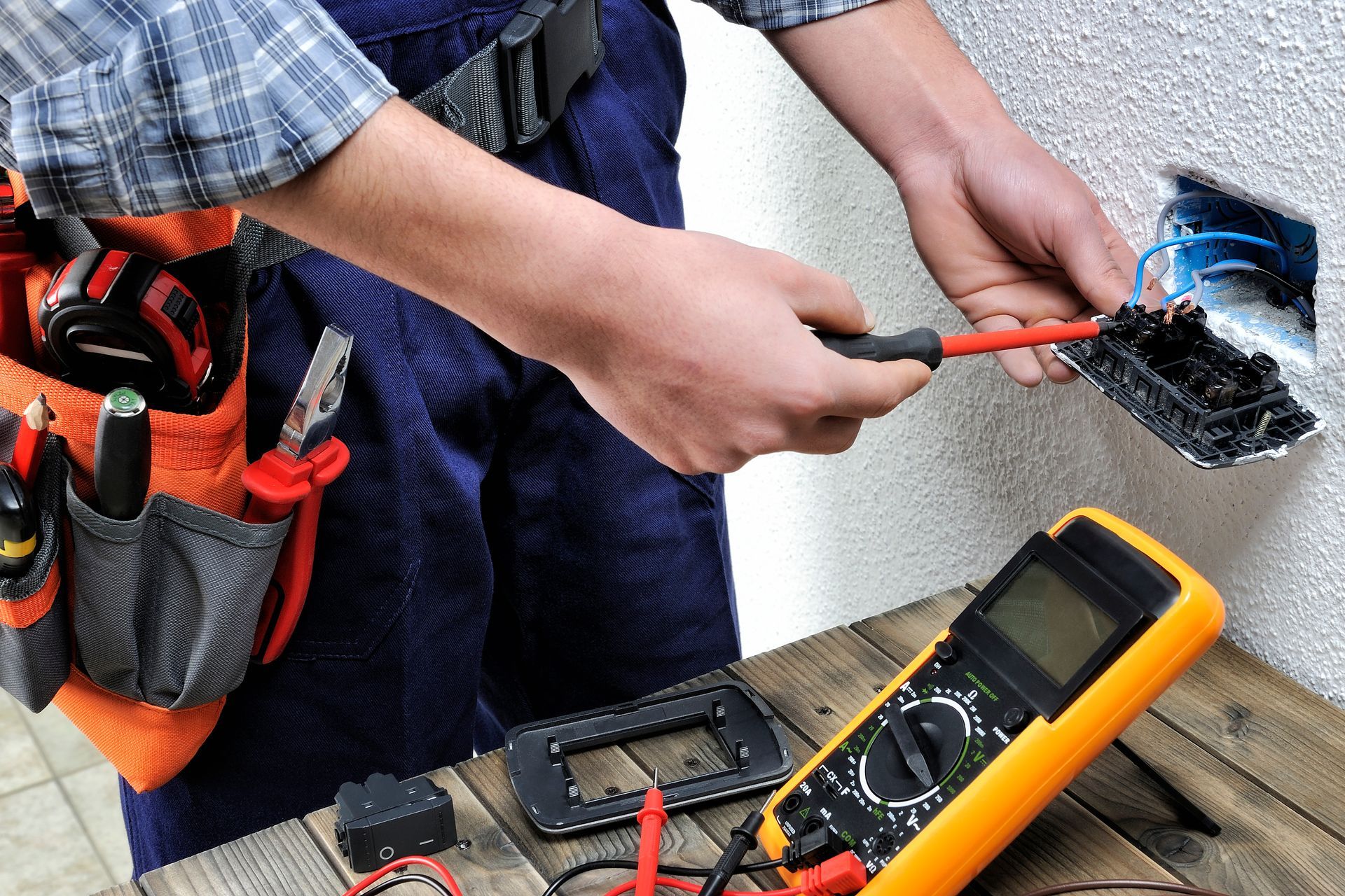 Person using tools to inspect electrical wiring on a workbench, with multimeter and tool belt nearby Person using tools to inspect electrical wiring on a workbench, with multimeter and tool belt nearby