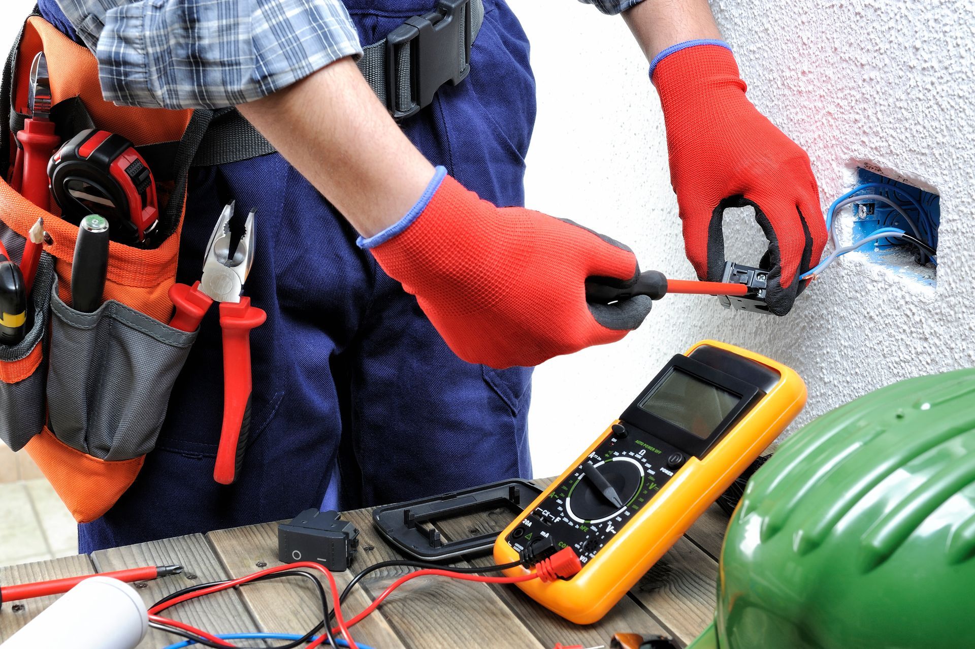 Electrician in red gloves testing wires with a multimeter near an outlet and tools on a workbench