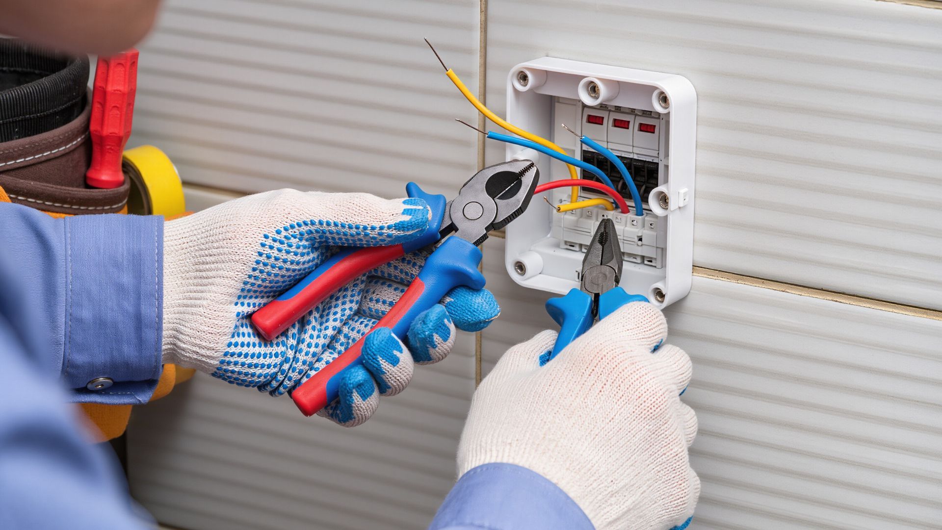 Gloved electrician wiring an outdoor electrical box with blue-handled pliers and exposed colored wires
