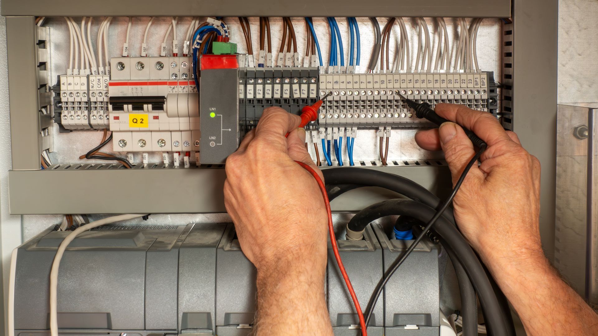 Technician wiring an electrical control panel with multimeter probes and organized colored cables.