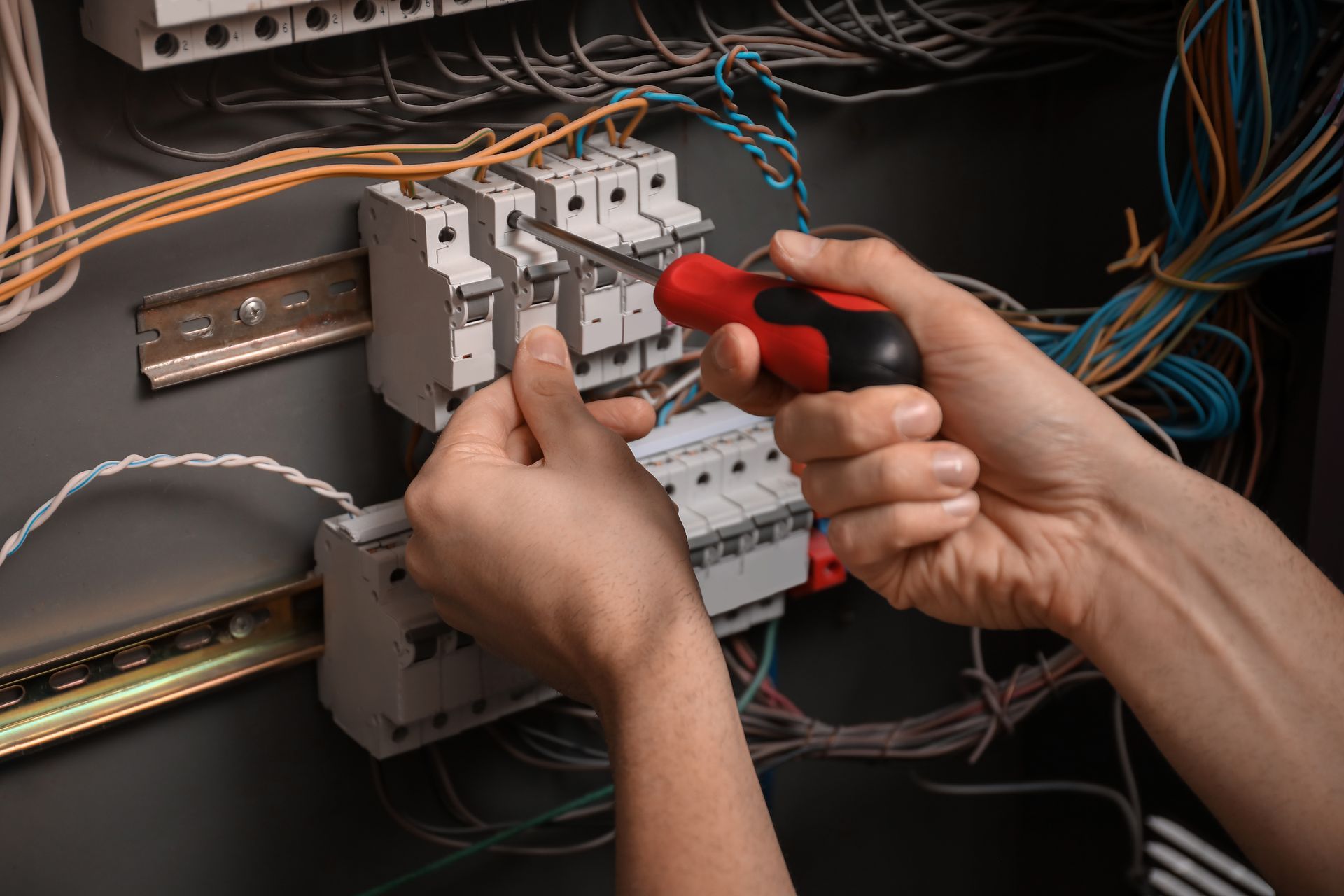 Hands wiring an electrical control panel with a red screwdriver among gray components and cables