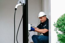 Technician in hard hat inspecting an electrical panel beside a doorway, outdoors near greenery Technician in hard hat inspecting an electrical panel beside a doorway, outdoors near greenery