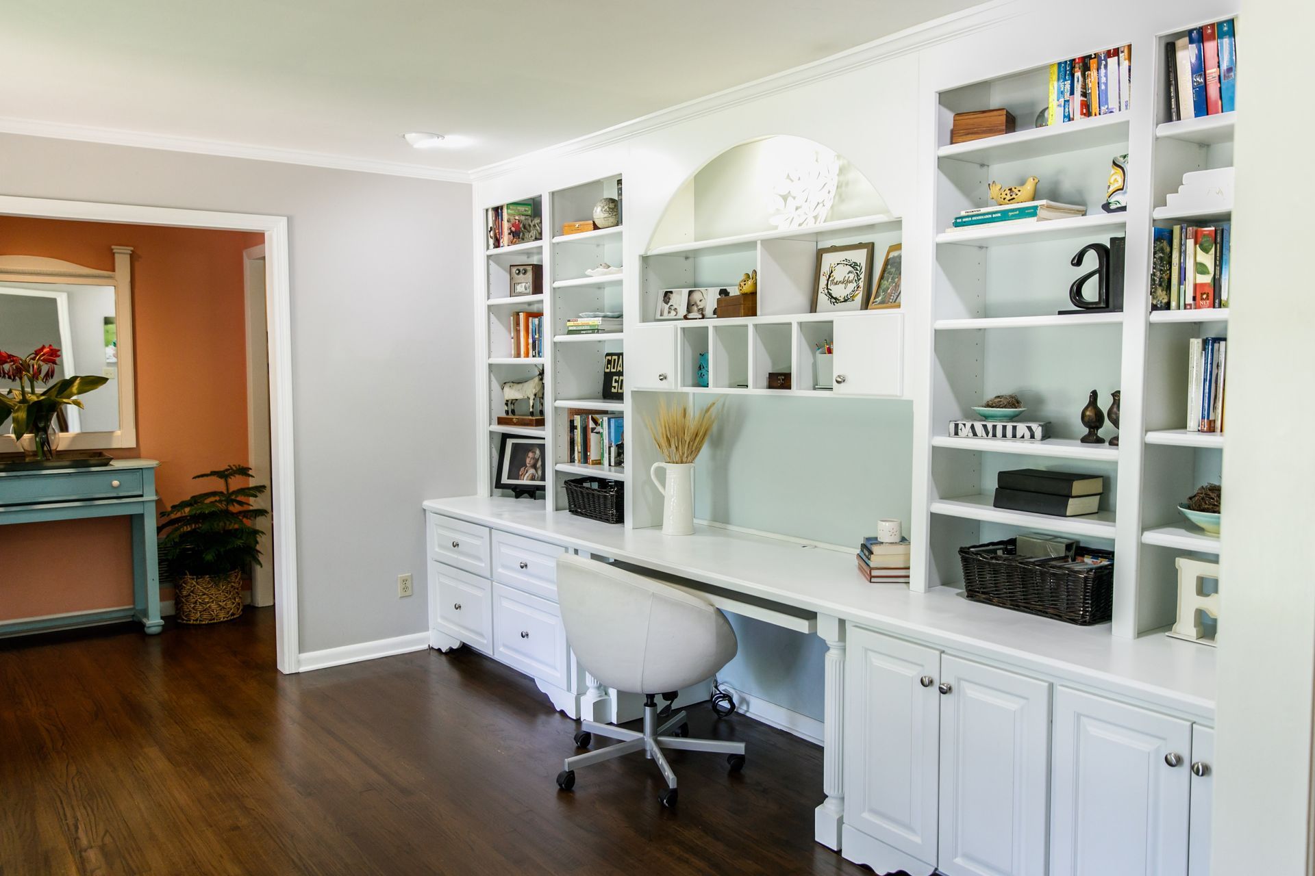 Built-in white desk with shelves and cabinets in a home office with dark wood floors.