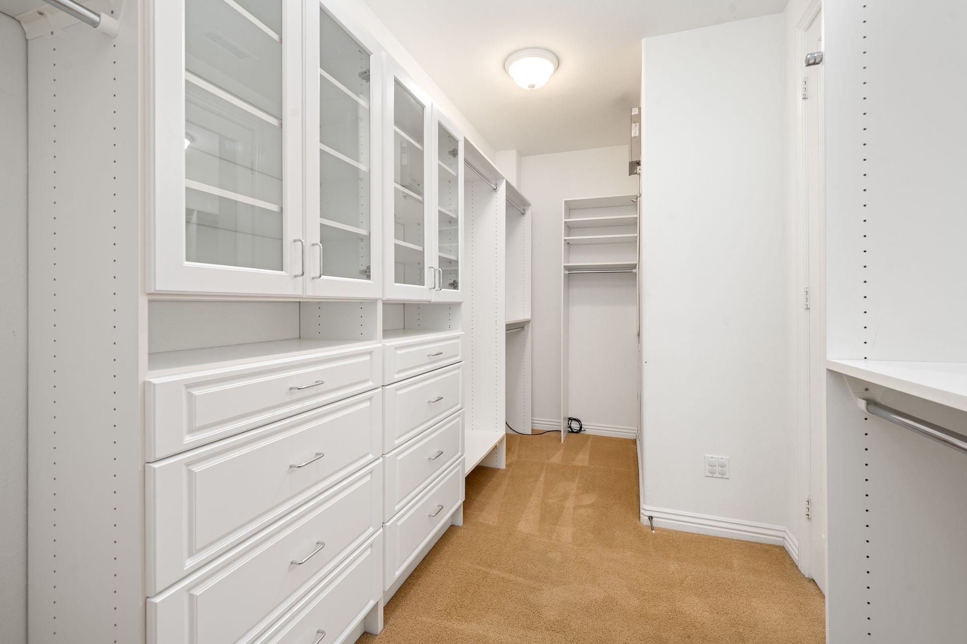White walk-in closet with built-in shelving and drawers; tan carpet, bright overhead lighting.