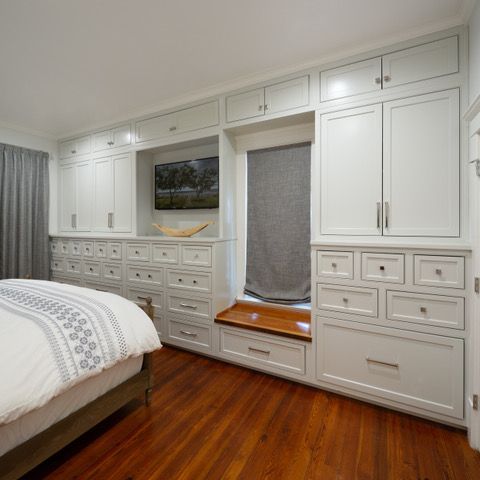 Built-in white bedroom storage with drawers and cabinets surrounding a window seat and TV on a hardwood floor.