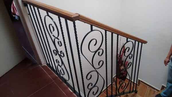 Escalera con pasamanos de madera y balaustres de hierro forjado negro; azulejos marrones en los escalones.