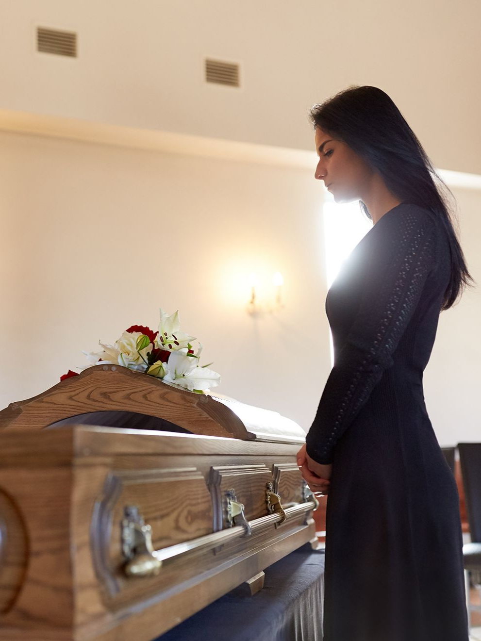 Woman in black dress at a funeral, facing a wooden casket with flowers.