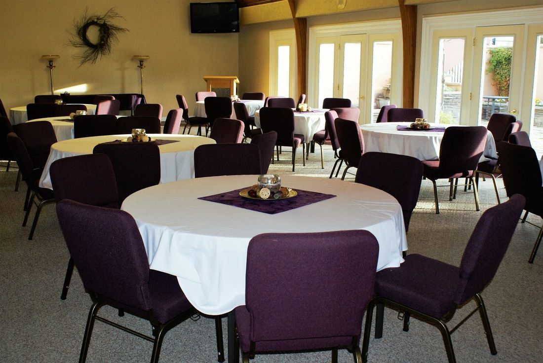 Banquet room with round tables, white tablecloths, and purple chairs by large windows.