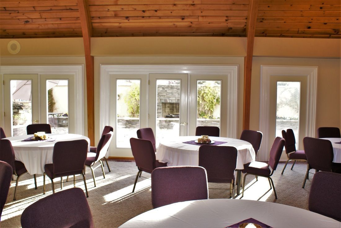 Sunlit dining room with round tables, chairs, and large windows under a wood-paneled ceiling