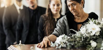 Woman leaning over a casket at a funeral, with white flowers and mourners in the background