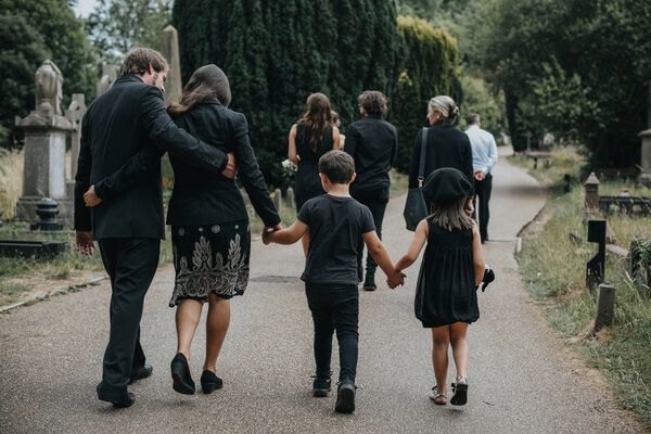 Group walking hand in hand down a tree-lined cemetery path beside headstones