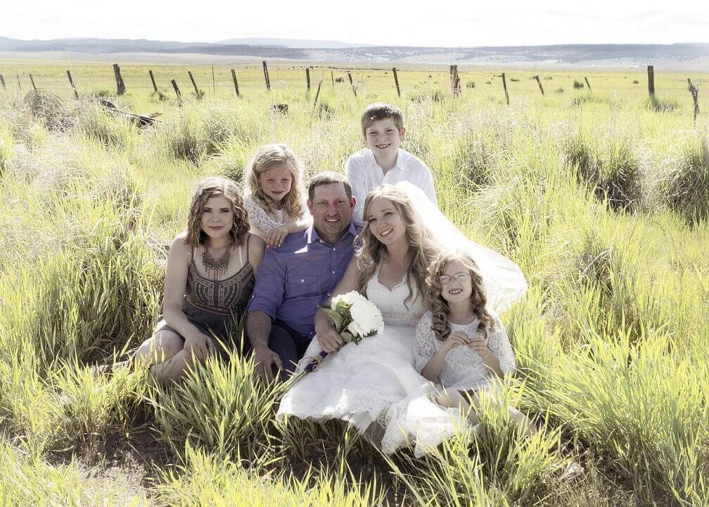 Family seated in tall grass outdoors, with a bride holding flowers in the center.
