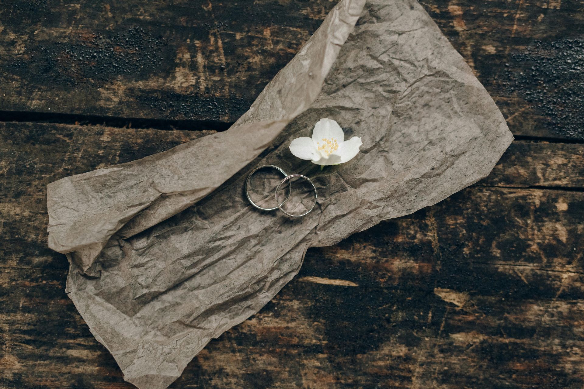 A pair of wedding rings and a flower are wrapped in brown paper on a wooden table.
