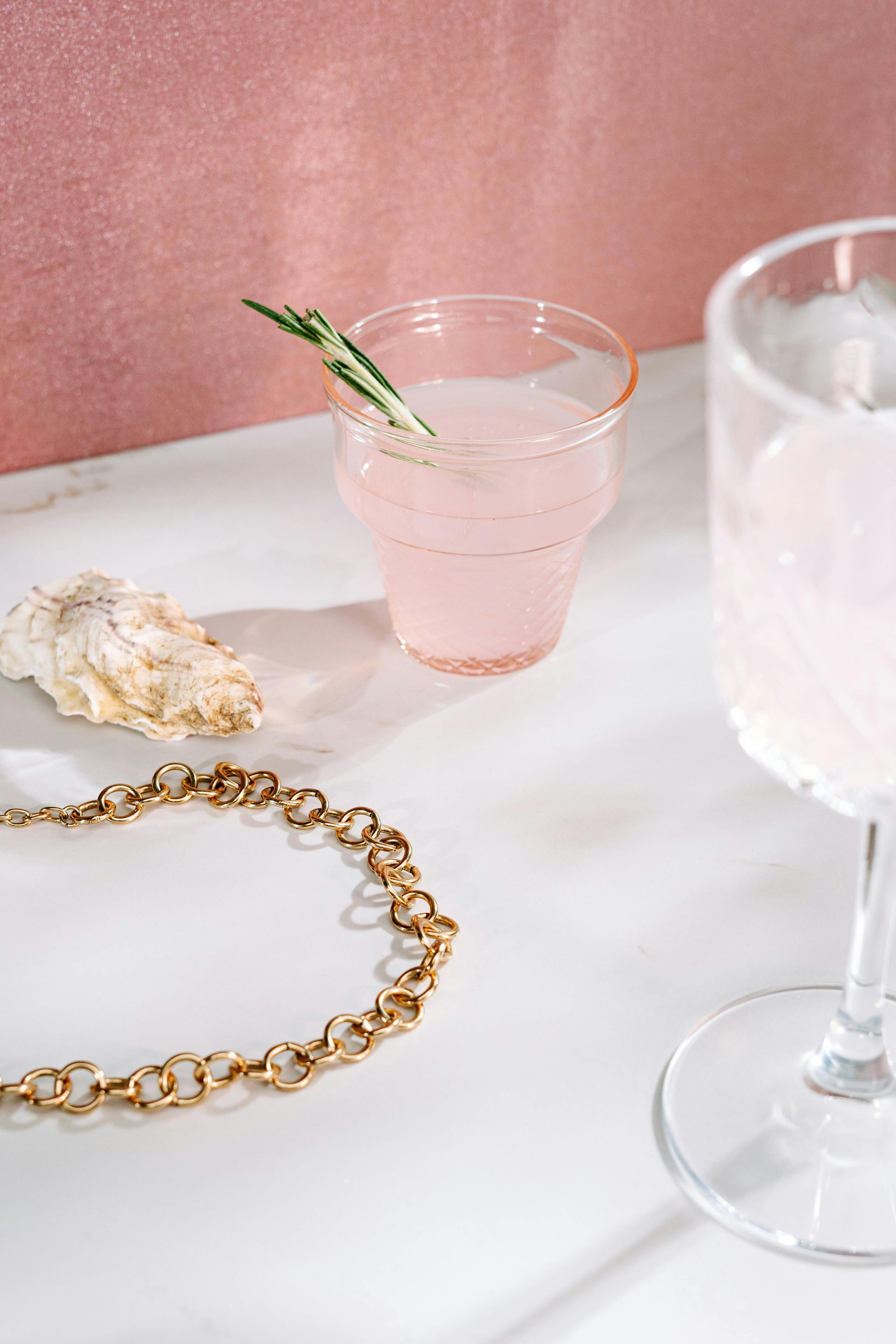 A glass of pink liquid is sitting on a table next to a necklace and a wine glass.