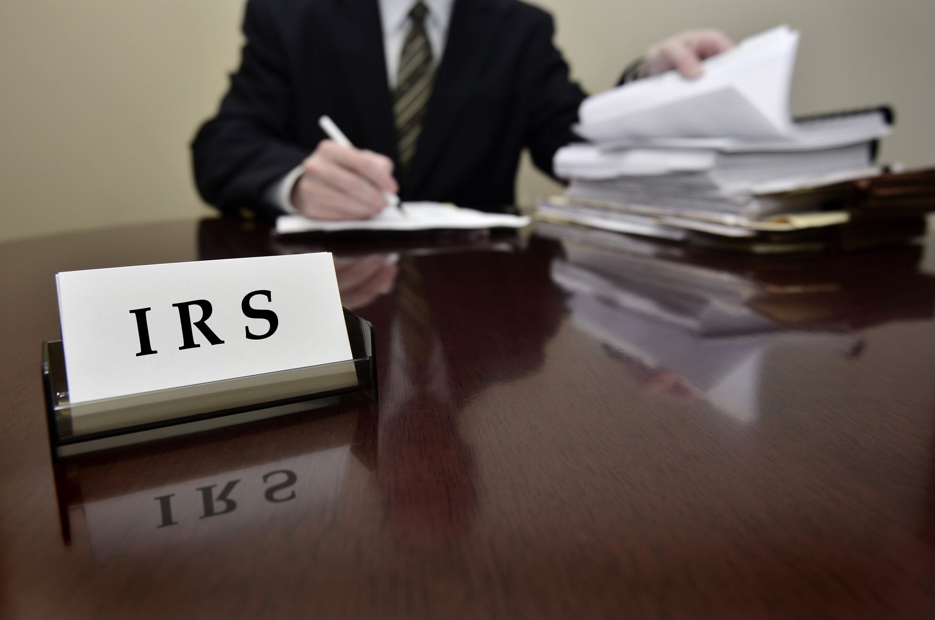 IRS desk sign beside a man in a suit writing at a desk with stacked papers