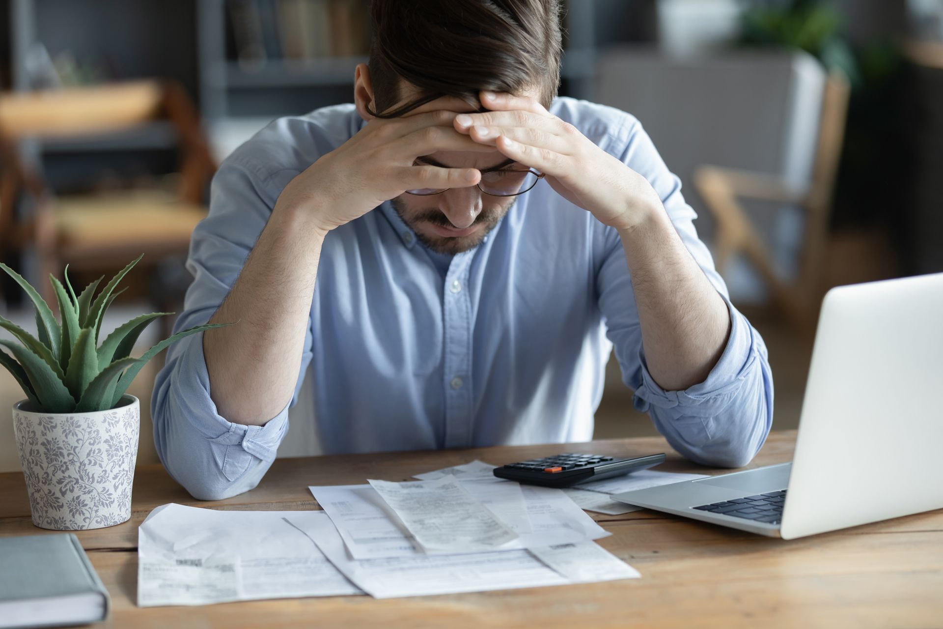Frustrated person at desk, head in hands, surrounded by papers and a laptop