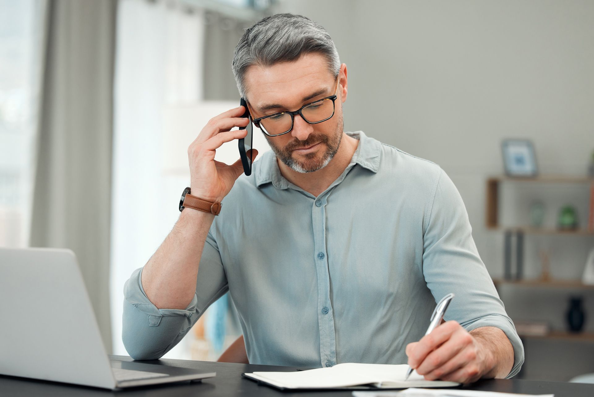 Man at desk on a phone call while writing notes beside an open laptop