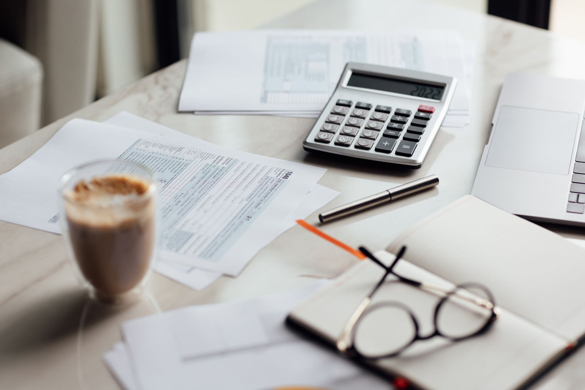 Desk with calculator, papers, coffee, pen, and eyeglasses in a bright office setting