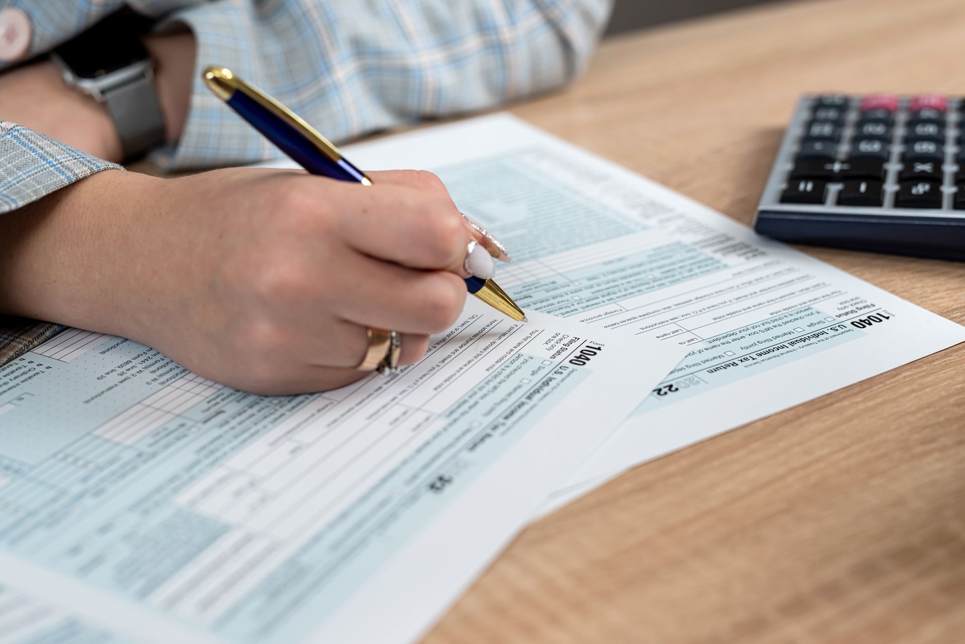 Hand writing on a form with a blue pen beside a calculator on a wooden desk