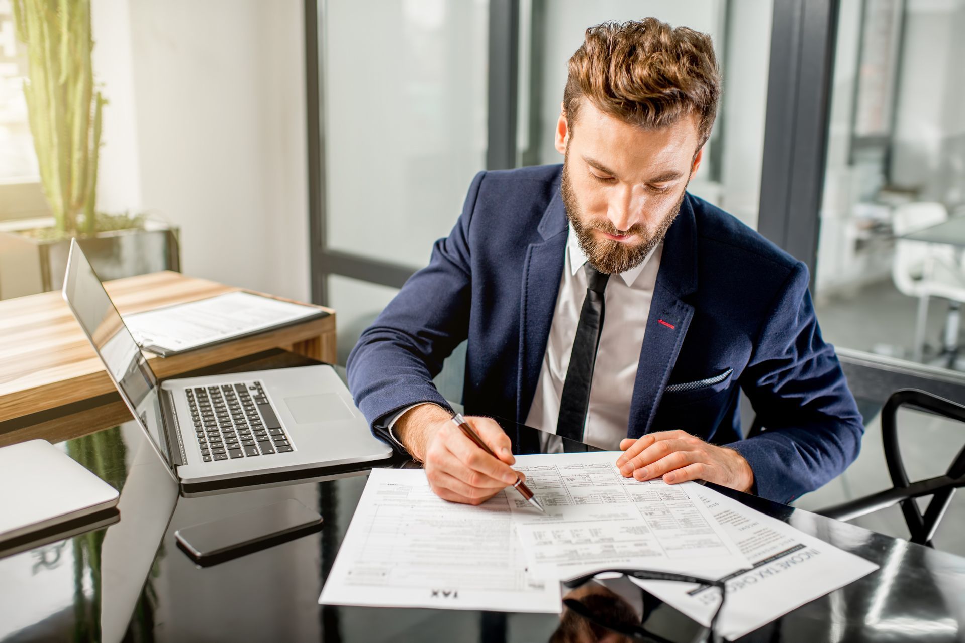 Businessman in a navy suit reviewing documents at a glass desk with a laptop nearby