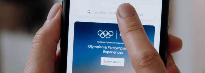 Mano tocando la pantalla de un teléfono que muestra el logotipo de los Juegos Olímpicos y texto relacionado con las experiencias.