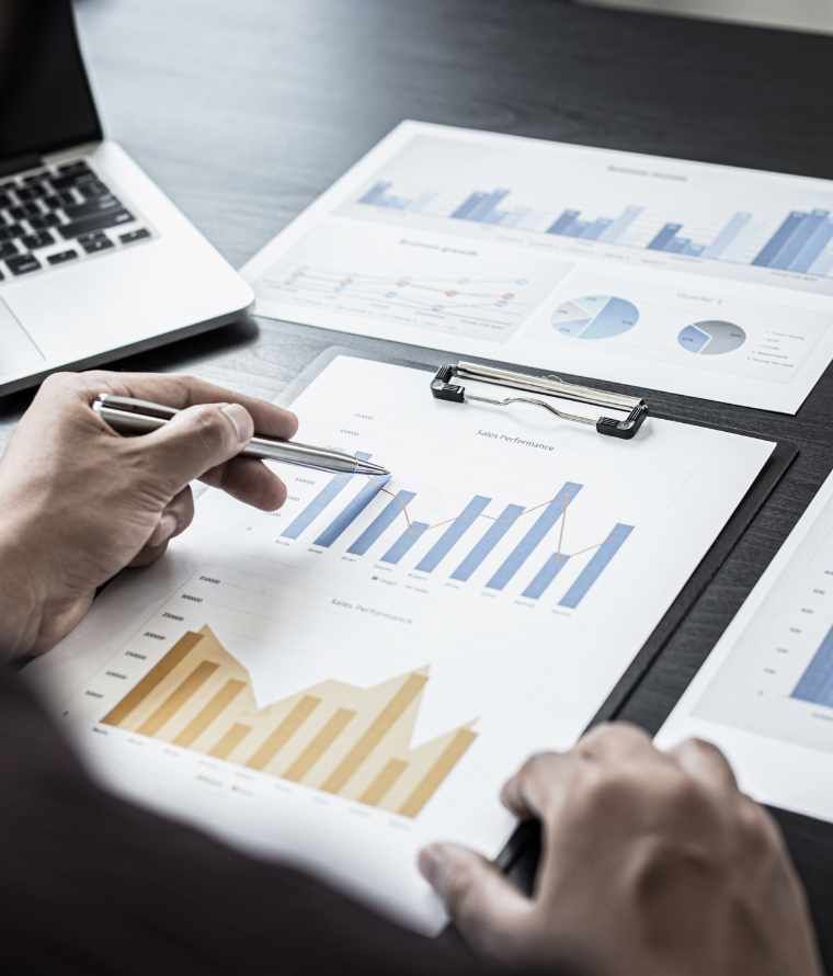 Persona analizando informes financieros con gráficos y una computadora portátil en un escritorio oscuro.