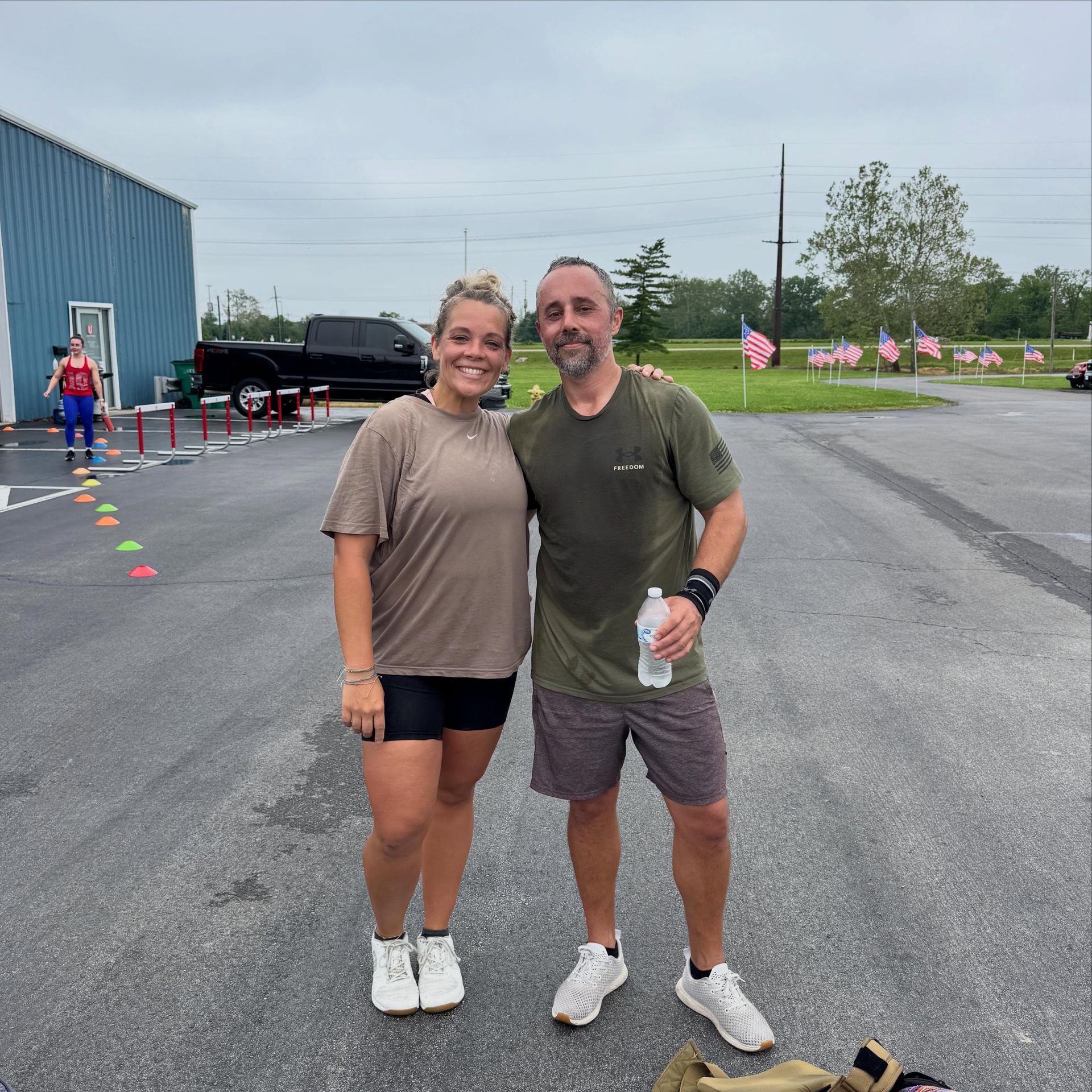 Two people standing arm-in-arm on a wet parking lot near a blue building and flag-lined road.