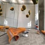 Modern lounge with orange sculptural chair, white tables, hanging decor, and gray flooring in a bright interior.