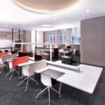 Modern open-plan office with white desks, black chairs, red accents, and large windows