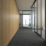 Empty modern hallway with wood-paneled wall, glass partition, and dark carpet leading to bright windows.