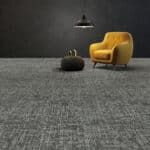 Gray carpeted room with a mustard yellow armchair, black hanging lamp, and round ottoman.