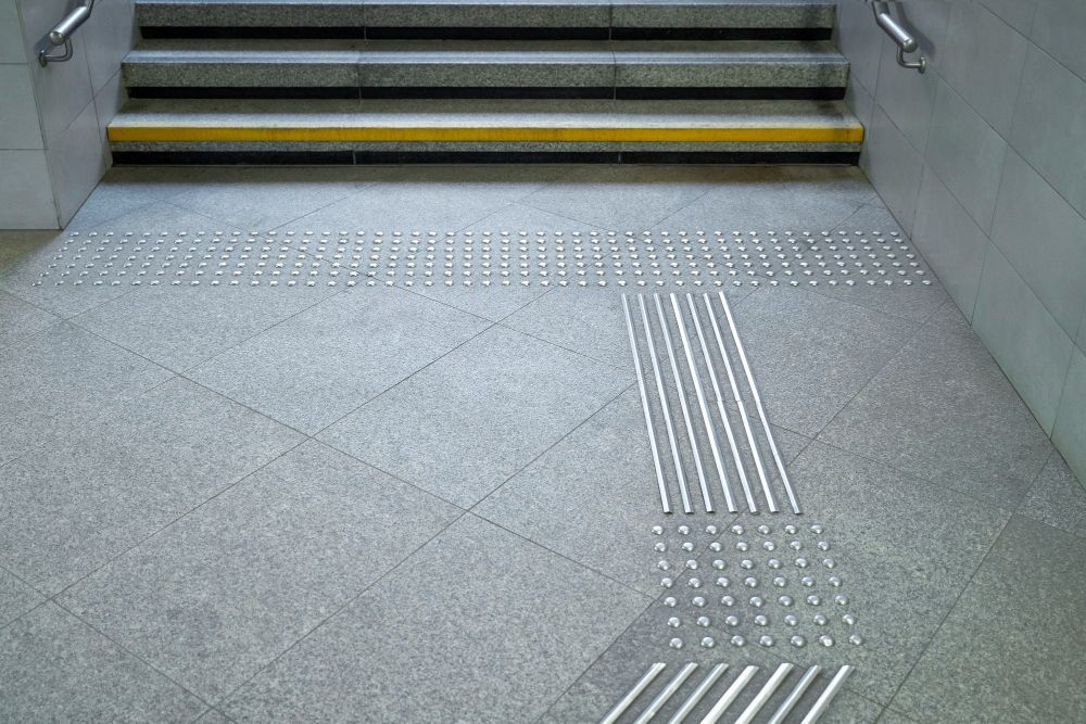 Tactile paving leads to a staircase with yellow-edged steps in a gray indoor corridor
