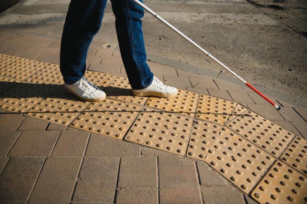 A person uses a white cane to navigate along a yellow tactile paving path on a sidewalk.