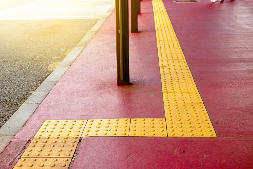 Yellow tactile paving on a red sidewalk near a road crossing, with black poles along the curb