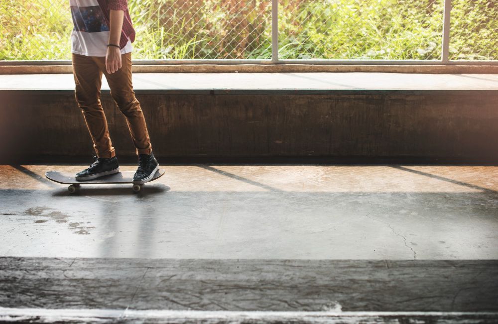 A person skateboarding on a smooth concrete surface in an indoor, sunlit space with a wire fence background.