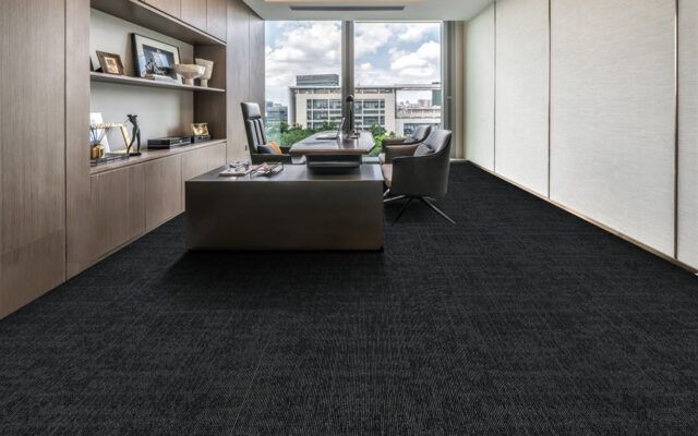 Modern office lounge with dark carpet, gray seating, built-in shelves, and large windows overlooking a city.