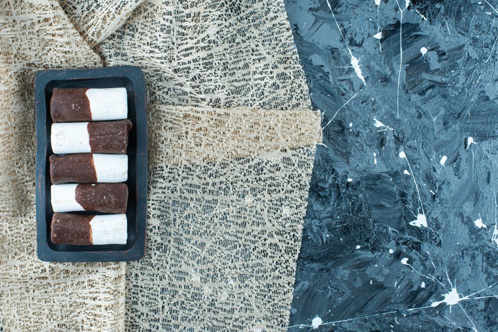 Five rectangular cookies, half-dipped in white frosting, arranged on a black tray atop a textured brown cloth.