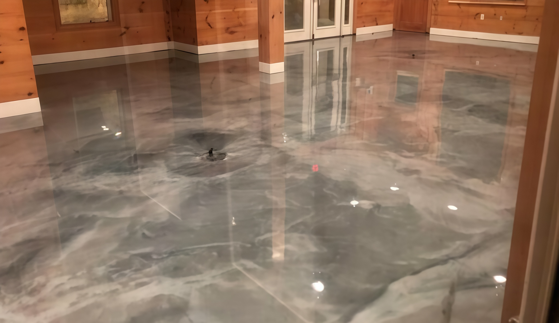 A person in work clothes uses a squeegee to spread smooth, glossy orange epoxy resin on a floor.
