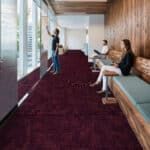 Airport terminal hallway with maroon carpet, people seated on benches, and a person standing near windows.