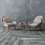 Two beige lounge chairs facing each other with a small round table on a gray textured floor and wall.