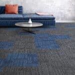 Modern lounge with blue sofa, orange pillow, round white coffee table, and patterned gray-blue carpet.