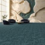 A small black object sits on a teal carpet in front of an abstract wall sculpture.