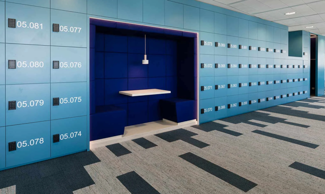 Blue locker room with numbered lockers and a central bench area beneath a hanging light.