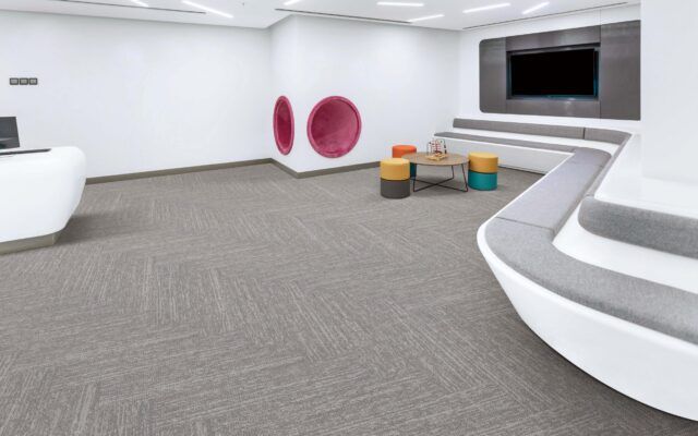 Modern white lounge with curved seating, gray carpet, colorful stools, and a wall-mounted TV